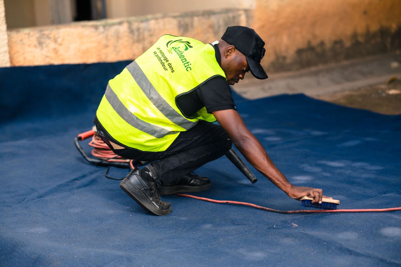 A worker dressed in a safety vest cleans a blue carpet outdoors with a brush.